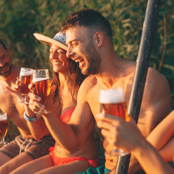 Happy group of young people drinking beer on a dock by the river during the summer sunny day